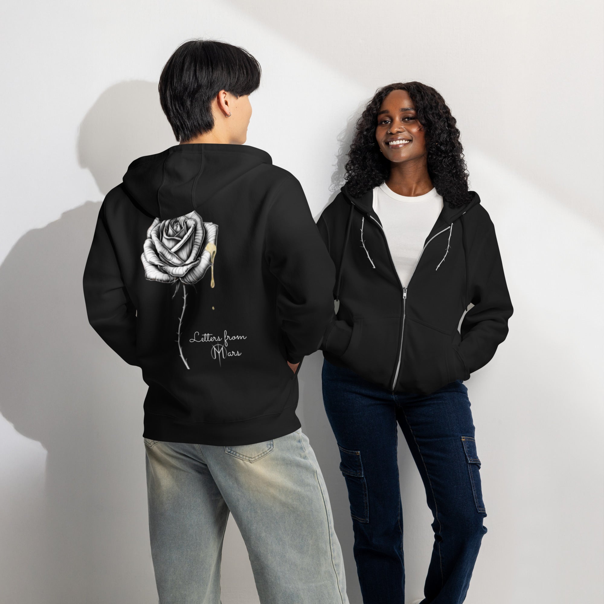 Two people wearing black hoodies with a Letters from Mars rose design on the back, standing against a white wall.